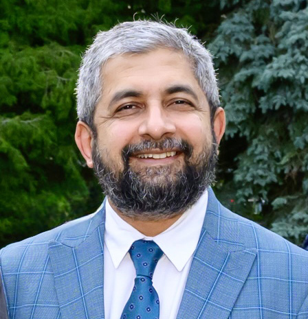 Dr. Iftikhar Chaudhry, wearing a blue checkered suit and tie, smiles warmly in an outdoor setting with green trees in the background, representing trusted and professional care – IC Laser Eye