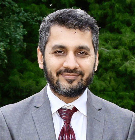 Dr. Imtiaz Chaudhry, dressed in a gray suit with a red tie, smiles confidently while standing outdoors with green trees in the background, representing expert and compassionate eye care – IC Laser Eye