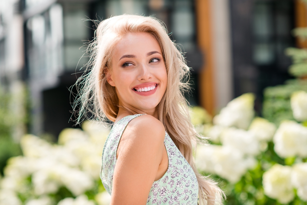 Smiling woman outdoors with bright flowers – The Pennsylvania Surgery and Laser Center A young woman with long blonde hair smiles radiantly while standing outdoors surrounded by blooming white flowers, conveying beauty, confidence, and natural elegance – The Pennsylvania Surgery and Laser Center